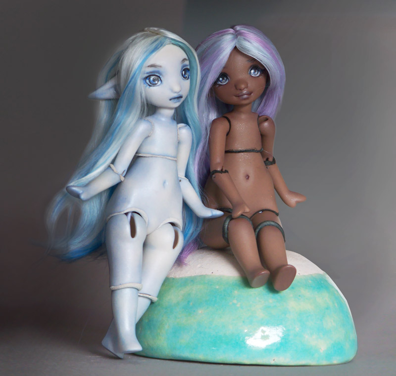 two tiny porcelain ball jointed elf dolls, one with blue and white hair and pale blue skin and the other with pastel blue and purple hair and dark skin.